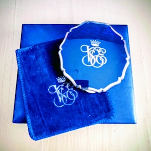 May include: A royal blue gift set featuring a box, a small square cloth, and a decorative tray. Each item has a white monogram design with a crown. The tray has a scalloped edge.