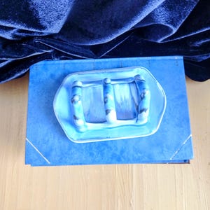 May include: A blue ceramic soap dish with three raised bars, resting on a blue book. The dish has a light blue and white marbled design. The background features a dark blue velvet fabric.