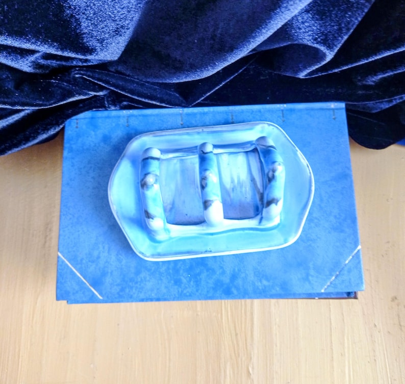 May include: A blue ceramic soap dish with three raised bars, resting on a blue book. The dish has a light blue and white marbled design. The background features a dark blue velvet fabric.