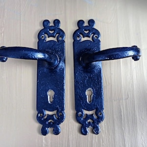 May include: Two ornate, navy blue door handles. Each handle has a decorative backplate with a keyhole and a curved handle. The design includes scrollwork and a stylized top. The handles are mounted on a light-colored surface.