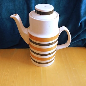 Puede incluir: Una tetera cilíndrica de cerámica con base blanca y rayas horizontales en tonos alternos de amarillo mostaza y marrón oscuro. La tetera incluye una tapa a juego con una banda marrón, un asa curva y un pico vertedor cónico.