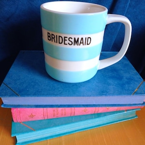 May include: A ceramic mug featuring horizontal light blue and white stripes with the word BRIDESMAID printed in black capital letters on the front. The mug has a white interior and a curved handle, positioned upright with a smooth, glossy finish.
