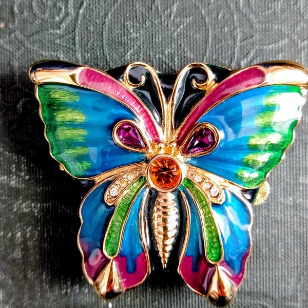 Könnte beinhalten: Eine farbenfrohe Schmetterlingsbrosche mit goldfarbenen Metallarbeiten. Die Flügel sind in Blau-, Grün-, Rosa- und Lilatönen bemalt, mit einem orangefarbenen Mittelstein und kleinen, klaren Kristallen. Die Brosche hat einen schwarzen Hintergrund.