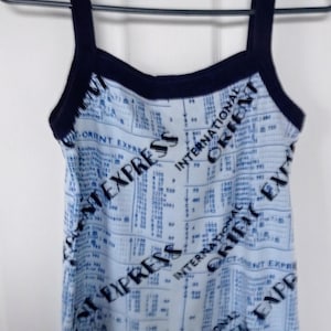 May include: A light blue tank top with a print of what appears to be a ledger or financial document. The words "INTERNATIONAL ORIENT EXPRESS" are printed diagonally across the front in black. The trim and straps are navy blue.
