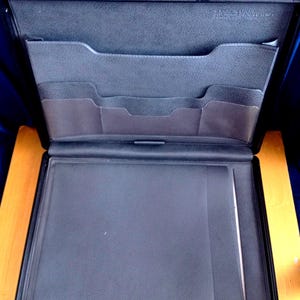 May include: A black leather-textured portfolio case with a hinged opening. The interior features a top section with multiple horizontal pockets for documents and a large bottom compartment with a vertical sleeve for holding papers or a notepad securely.