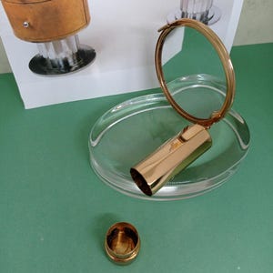 Op de afbeelding: Een goudkleurige compacte spiegel met een ovale basis van helder glas. De spiegel is open en toont het reflecterende oppervlak. Een kleine, goudkleurige container staat in de buurt op een groene ondergrond. Een bureau staat op de achtergrond.