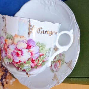 Könnte beinhalten: Weißes Tee-Set mit Blumendesign. Die Teetasse hat einen Henkel und das Wort "Carrgum" in goldfarbenen Buchstaben. Das Blumendesign umfasst rosa, lila und orangefarbene Blumen mit grünen Blättern.