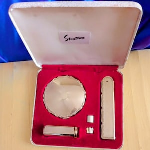 May include: An open vintage Stratton compact set. The set includes a round compact, lipstick case, and other accessories, all in a silver-tone finish. The interior is lined with red velvet, and the case is white with the name "Stratton" on the lid.