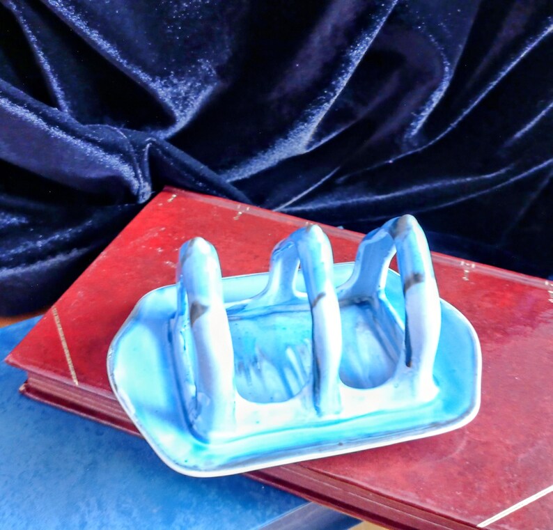 May include: A blue ceramic toast rack with three arched slots, resting on a red book. The rack has a rectangular base and is painted with shades of blue and white. The background features a dark blue velvet fabric.
