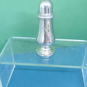 May include: A silver salt shaker with a rounded top and a decorative emblem. The shaker has a classic design and is made of a shiny, reflective metal. The salt shaker is approximately 4 inches tall.
