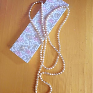 May include: A cream-coloured pearl necklace draped over a patterned box. The box has a white background with a pink and gold floral design. The necklace is on a yellow surface.