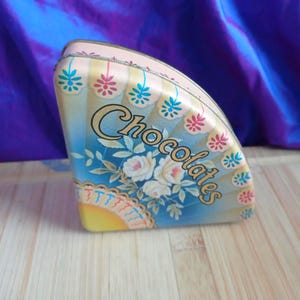 Puede incluir: Una caja de lata vintage en forma de abanico con la palabra "Chocolates" en escritura elegante. La caja presenta un fondo azul claro con rosas blancas y un diseño de abanico decorativo. La parte superior y los bordes son de color rosa pálido.