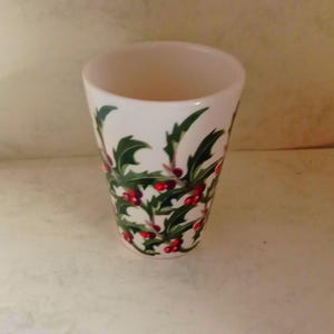 May include: A white ceramic tumbler decorated with green holly leaves and red berries. The tumbler has a cylindrical shape and a smooth, glossy finish. The holly design is repeated around the exterior, creating a festive holiday theme.