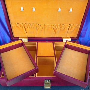 May include: Open burgundy jewelry box with a mustard yellow interior. The box has multiple compartments and trays, with a gold-colored clasp. The interior is lined with a soft, textured fabric. The box is open, revealing its storage space.