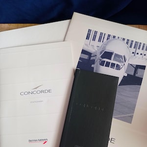 以下が含まれることがあります： ConcordeのロゴとBritish Airwaysのブランド名が入った白いステーショナリーフォルダーと、Concordeの文字がエンボス加工された黒いノート。別の白いシートには、Concorde航空機の機首の白黒写真が印刷されています。