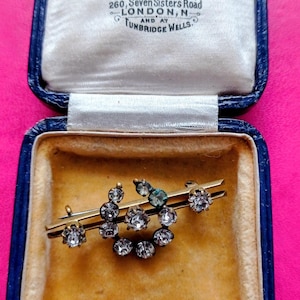 May include: A gold-toned bar brooch featuring a circular arrangement of clear, faceted stones, housed in a dark blue velvet-lined jewellery box. The interior lid displays the text: E & R. S. HARRISON JEWELLERS & CO. 260, SEVEN SISTERS ROAD LONDON, N AND AT TUNBRIDGE WELLS.