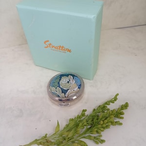 以下が含まれることがあります： 青、緑、白の花柄が施されたシルバーのコンパクトと、"Stratton"の文字がプリントされた水色の箱。緑の葉の小枝が前景にあります。