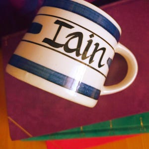 May include: White ceramic mug with blue stripes and the name "Iain" in black lettering. The mug has a curved handle and is resting on a stack of books with red, green, and purple covers. The mug is a personalized gift.