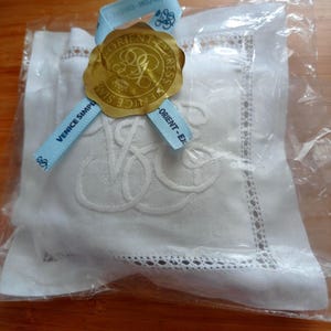 Könnte beinhalten: Ein quadratisches Säckchen aus weißem Leinen mit gesticktem Monogramm, verpackt in transparenter Folie. Ein hellblaues Band ist oben mit einem goldenen Wachssiegel befestigt, das den Schriftzug Venice Simplon-Orient-Express trägt.
