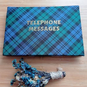 May include: A rectangular notebook with a blue, green, and black plaid pattern. The gold text on the cover reads "TELEPHONE MESSAGES." A small bundle of dried blue flowers tied with twine rests below the notebook.