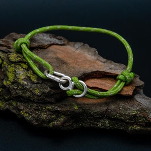 Könnte beinhalten: Ein grün-weißes Seilarmband mit Silberverschluss. Das Armband ist geknotet und liegt auf einem Stück strukturierter, dunkelbrauner Rinde. Der Hintergrund ist dunkelblau.