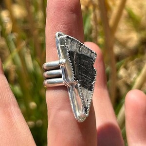 Può includere: Anello d'argento con una grande pietra nera di forma irregolare e superficie strutturata. La pietra è incastonata in una lunetta d'argento con un bordo decorativo. L'anello ha un design a tripla fascia.