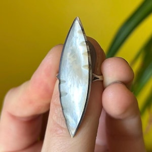 May include: A silver ring featuring a large, elongated, pearlescent stone. The stone has a subtle iridescent sheen and is set in a silver band. The ring is held against a yellow background.