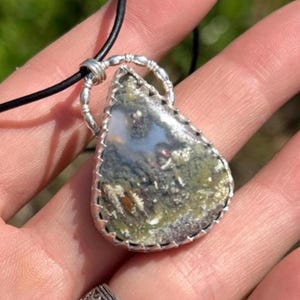 May include: A teardrop-shaped pendant necklace featuring a natural stone with green, white, and brown inclusions. The stone is set in a silver-colored metal frame and hangs from a black cord.