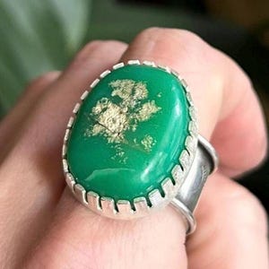 May include: A silver ring featuring a large, oval-shaped green stone with a textured, metallic gold-colored inclusion in the center. The stone is set in a serrated silver bezel, and the band is made of a smooth, polished silver metal.