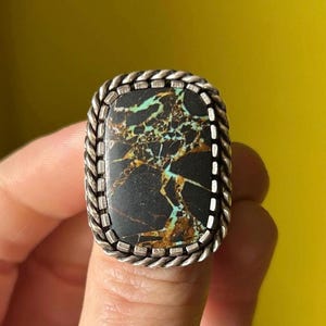 May include: A silver ring with a large, rectangular black stone featuring a unique pattern of brown, turquoise, and gold veining. The stone is set within a braided silver border, creating a striking contrast.