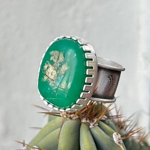 May include: A silver ring with a green gemstone set in a scalloped bezel. The gemstone has a unique pattern of gold flecks throughout.