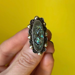 Peut inclure: Une bague ovale avec une pierre centrale en coquillage d'ormeau irisé. La bague a un sertissage argenté avec des accents dorés sur les bords. La coquille d'ormeau affiche des nuances de vert, de bleu et de violet. La bague est sur fond jaune.