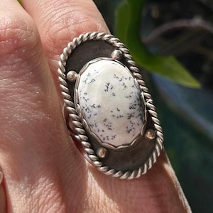 May include: An oval ring featuring a white dendritic opal stone with black fern-like inclusions. The stone is set in a dark metal bezel surrounded by a twisted rope border and accented with four small silver spheres at the top and bottom edges.