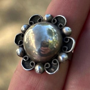 May include: A silver ring featuring a large, smooth, polished circular center dome. The dome is surrounded by a decorative border of alternating small silver spheres and delicate, curved wire scrollwork, creating a flower-like appearance.