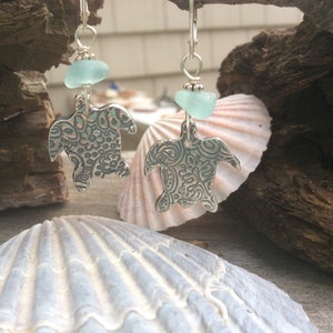 Könnte beinhalten: Silberne Schildkrötenohrringe mit einem hellblauen Meerglasschmuckstück. Die Schildkröten haben ein detailliertes Blumenmuster.