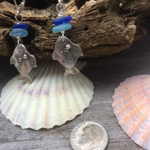 Könnte beinhalten: Silberne Fischohrringe mit blauen und hellblauen Glasperlen, die an der Unterseite hängen. Die Ohrringe werden auf einem Stück Treibholz mit Muscheln präsentiert.