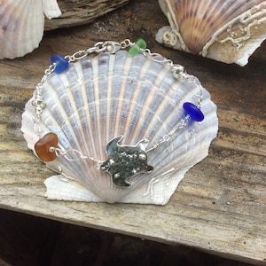 Puede incluir: Un brazalete de plata con un dije de tortuga y cuentas de vidrio de mar en tonos azules, verdes y marrones. El brazalete se muestra sobre una concha.