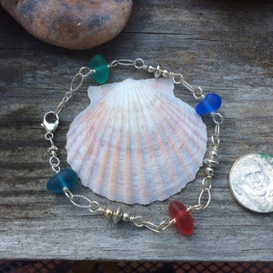 May include: A silver chain bracelet with four sea glass beads in shades of blue and red. The bracelet is displayed on a seashell and a UK 20 pence coin.