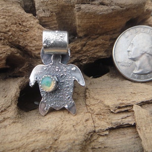 Puede incluir: Un colgante de plata en forma de tortuga con una piedra de ópalo verde engastada en el centro. El colgante es texturizado y tiene un aspecto rústico. Es aproximadamente del tamaño de una moneda de 25 centavos.