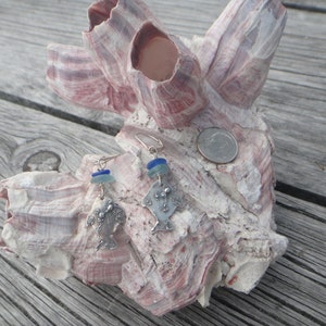 Könnte beinhalten: Ein Paar silberne Ohrringe mit blauen und grünen Glasperlen und einem silbernen Fisch-Anhänger. Die Ohrringe werden auf einer Muschel präsentiert.