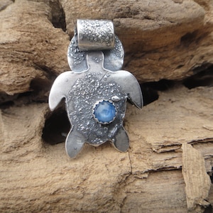 Puede incluir: Pendiente de plata en forma de tortuga con una piedra preciosa azul engastada en el centro. La tortuga tiene una superficie texturizada y un bucle para sujetar una cadena.