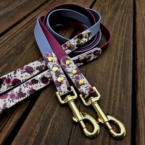 May include: A multi-colored dog leash featuring a combination of solid light blue and deep magenta straps. The leash includes sections with a white floral pattern displaying dark purple and pink roses, secured by gold-toned metal rivets and two gold swivel clips.