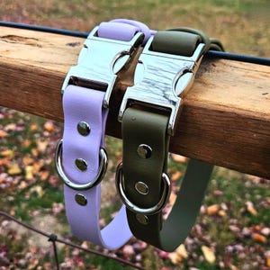May include: Two pet collars, one in light lavender and one in olive green, each featuring a silver-toned metal side-release buckle, a matching silver D-ring for leash attachment, and three silver-toned metal rivets securing the strap.