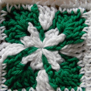 May include: A close-up of a crocheted square with a white and green pattern. The square is made of yarn and has a textured surface.