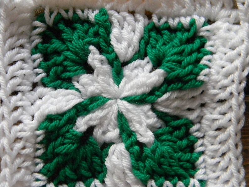 May include: A close-up of a crocheted square with a white and green pattern. The square is made of yarn and has a textured surface.
