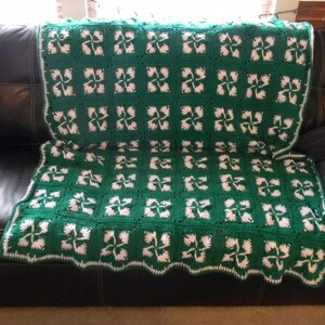 May include: A green and white crocheted throw blanket with a four-leaf clover pattern. The blanket is draped over a black leather couch.