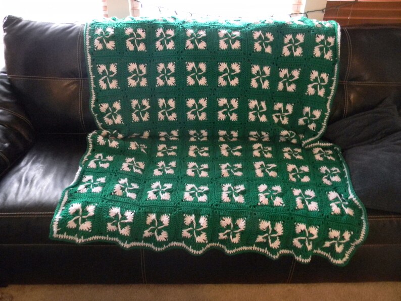 May include: A green and white crocheted throw blanket with a four-leaf clover pattern. The blanket is draped over a black leather couch.