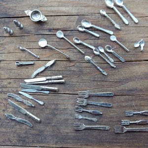 May include: A collection of miniature silver-colored cutlery, including spoons, forks, and knives, arranged on a rustic wooden surface. The set includes various sizes and styles of utensils, perfect for dollhouses or craft projects.