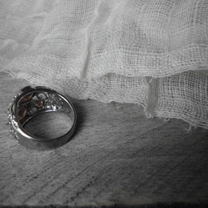 K&ouml;nnte beinhalten: Ein silberner Ring mit einem dekorativen Design mit mehreren kleinen Steinen. Der Ring liegt auf einer Holzoberfl&auml;che mit einem wei&szlig;en Stoff dar&uuml;ber drapiert.