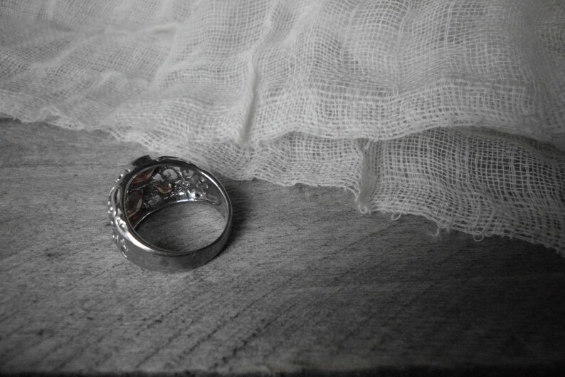 K&ouml;nnte beinhalten: Ein silberner Ring mit einem dekorativen Design mit mehreren kleinen Steinen. Der Ring liegt auf einer Holzoberfl&auml;che mit einem wei&szlig;en Stoff dar&uuml;ber drapiert.